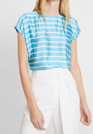 Vrouw draagt een lichtblauw-wit gestreept shirt met korte mouwen, gestopt in een hoge, witte broek met een stoffen riem.