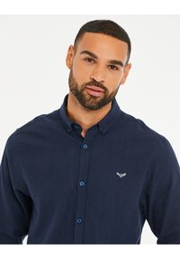 Camisa de botones azul marino hecha de tejido texturizado, con botones azules en contraste y un pequeño detalle bordado en el pecho.