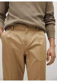 Karamelkleurige broeken met een rechte pasvorm, voorzien van een knoopsluiting, lussen voor een riem en een gladde stofstructuur. Grijze, ribbelige sweater is gedeeltelijk zichtbaar.