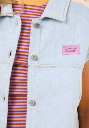 Veste en denim bleu clair avec une étiquette Oxbow violette, dotée de boutons argentés et d'un patch visible. En dessous, une chemise rayée en orange et violet.