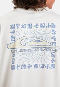 Hvid t-shirt med et blåt grafisk design med bølgelogo, cirkulær tekst og abstrakte mønstre på ryggen. Korte ærmer, afslappet pasform.