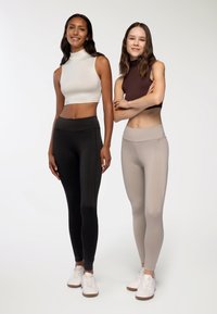 Dwie kobiety stoją obok siebie, ubrane w dopasowane ubrania sportowe. Po lewej: biały top bez rękawów i czarne legginsy. Po prawej: brązowy top bez rękawów i beżowe legginsy.