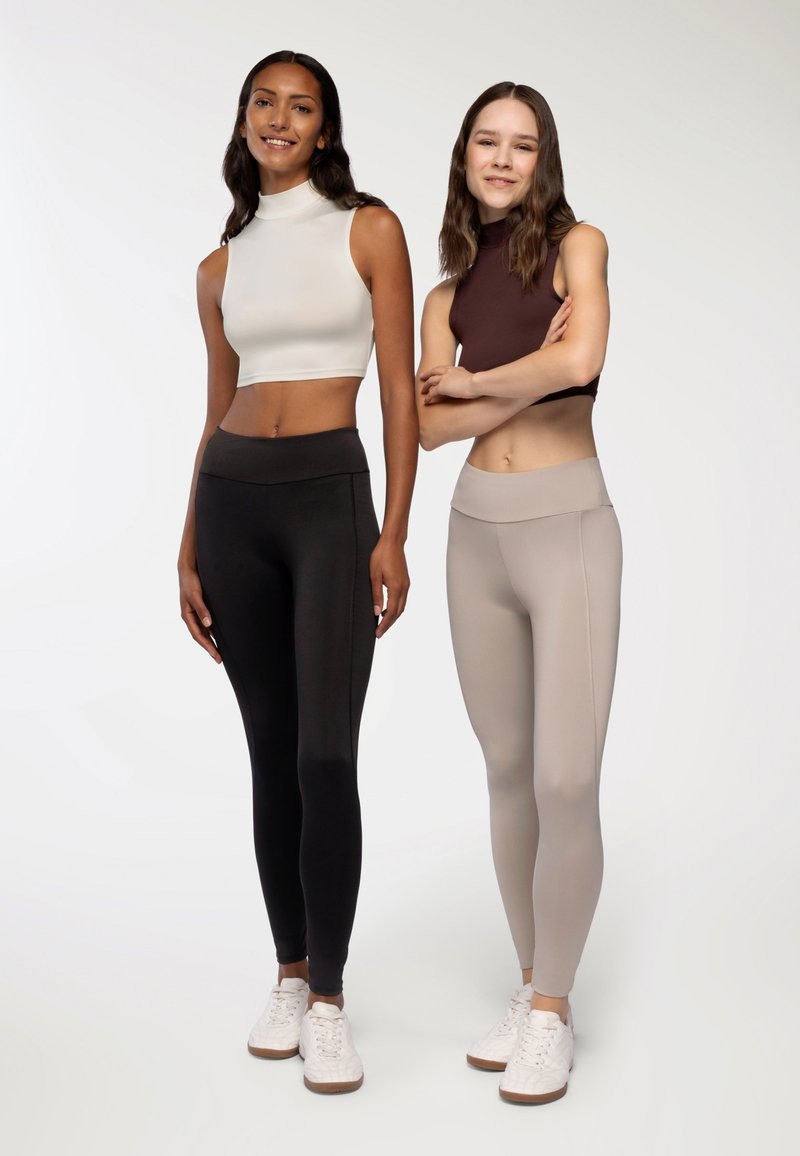 Dwie kobiety stoją obok siebie, ubrane w dopasowane ubrania sportowe. Po lewej: biały top bez rękawów i czarne legginsy. Po prawej: brązowy top bez rękawów i beżowe legginsy.