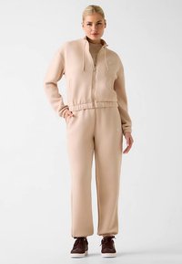 Beiger Kapuzenpullover mit Reißverschluss und passende Sweatpants aus weichem Stoff, mit tailliertem Bund und Fronttaschen. Braune Sneakers runden das Outfit ab.