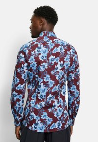 Bloemige blouse met een bordeauxrode basis, blauwe bloemen en groene stelen, lange mouwen, button-down kraag en een getailleerde pasvorm.