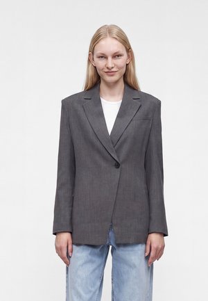 Jonge vrouw met lang blond haar, gekleed in een donkergrijze blazer, wit shirt en lichtblauwe jeans, staand tegen een effen achtergrond.