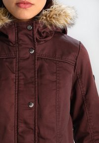 Bordowa kurtka z kapturem wyłożonym sztucznym futerkiem, zatrzaskami oraz gładką tkaniną z przeszyciami na przodzie.