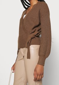 Cardigan marron tricoté avec un col en V et un design portefeuille, doté d'une texture côtelée et d'une ceinture latérale. Associé à un pantalon beige ajusté.