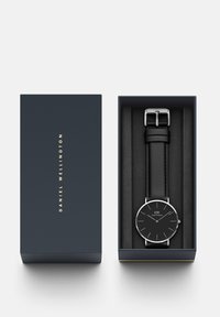 Black wristwatch with silver accents, enclosed in a navy box with gold text. Leather strap and minimalist round face featuring hour markers.