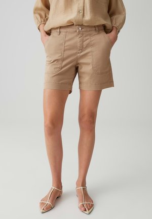 Woman wearing beige high-waist shorts with front patch pockets, beige long sleeve shirt, and white strappy pointed-toe sandals, hands in pockets.