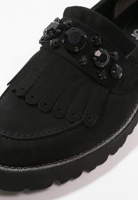 Svart sko med en mockaliknande ovandel, prydd med fransar och dekorativa svarta ädelstenar. Gummisula med en diskret skimrande kant.