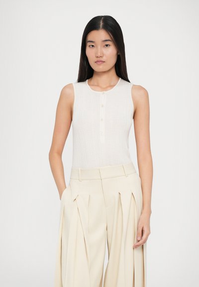 Vaaleanharmaa ilman hihoja oleva toppi, jossa on uritettu pinta ja nappiyksityiskohta edessä, yhdistettynä löysiin, kermavärisiin laskostettuihin housuihin.