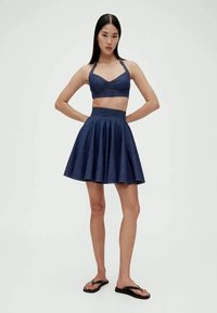 Mulher vestindo um top cropped halter azul-marinho, saia pregueada a condizer e chinelos pretos, de pé com as mãos na anca contra um fundo branco.
