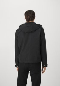 Svart jakke med hette, laget av glatt stoff. Har trykknapplommer og en enkel, strømlinjeformet design uten ekstra detaljer.