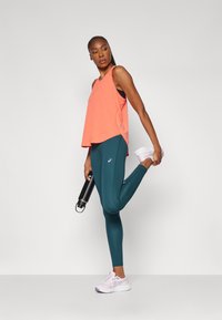 Korallfarget ermeløs treningstopp, teal leggings og lyse lilla joggesko. Modellen holder en svart vannflaske og strekker ut det ene beinet.