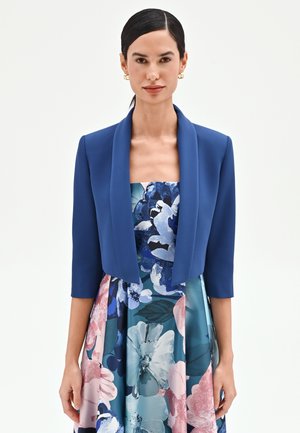Femme aux cheveux noirs lisses portant des boucles d'oreilles créoles dorées, un blazer court bleu roi et une robe bustier fleurie aux motifs bleus, roses et blancs.