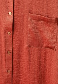 Rust-colored shirt fabric close-up showing a left chest pocket and a vertical row of four brown buttons.