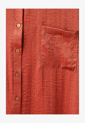 Primo piano di un tessuto di camicia color ruggine che mostra una tasca sul petto sinistro e una fila verticale di quattro bottoni marroni.