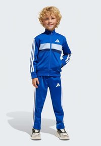 Blonds bērns smaida, ģērbies zilā Adidas sporta tērpā ar baltām svītrām un logotipu, stāv ar vienu roku kabatā un baltos sporta apavos.