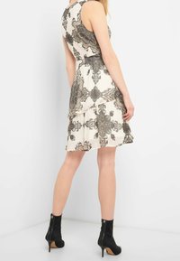 Ärmelloses Kleid in Creme mit schwarzem Blumenmuster. Verfügt über einen gestuften Rock und ein strukturiertes Oberteil. Kombiniert mit schwarzen Knöchelstiefeln.
