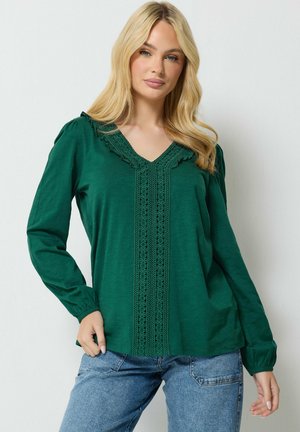 Mujer rubia con blusa de manga larga de color verde oscuro con detalles de encaje en la parte delantera y vaqueros azules, de pie contra una pared lisa y clara.