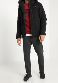 Chaqueta negra aislante con capucha, combinada con un sudadera roja y pantalones de chándal grises. Las zapatillas completan el conjunto, enfatizando la comodidad y la practicidad.