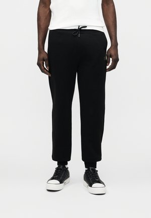 Mężczyzna ubrany w czarne spodnie ze ściągaczem i czarne sneakersy za kostkę z białymi podeszwami, stojący na tle jednolitego białego tła.