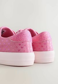 Des baskets roses pailletées avec de petits motifs de cœur et des semelles blanches épaisses, montrées de dos sur un fond neutre.