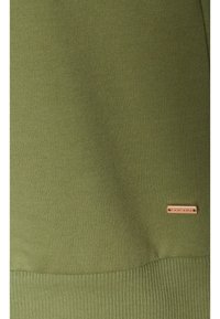 Olivgrüner gerippter Stoff mit glatter Textur und horizontalem Strickmuster. Verfügt über ein kleines metallisches Logo-Tag in der unteren rechten Ecke.