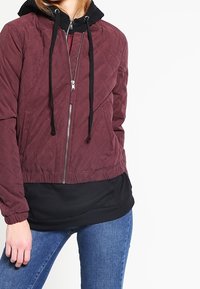Giacca trapuntata bordeaux con cappuccio nero, chiusura frontale con zip e polsini elasticizzati. Indossata con un felpa nera sotto.