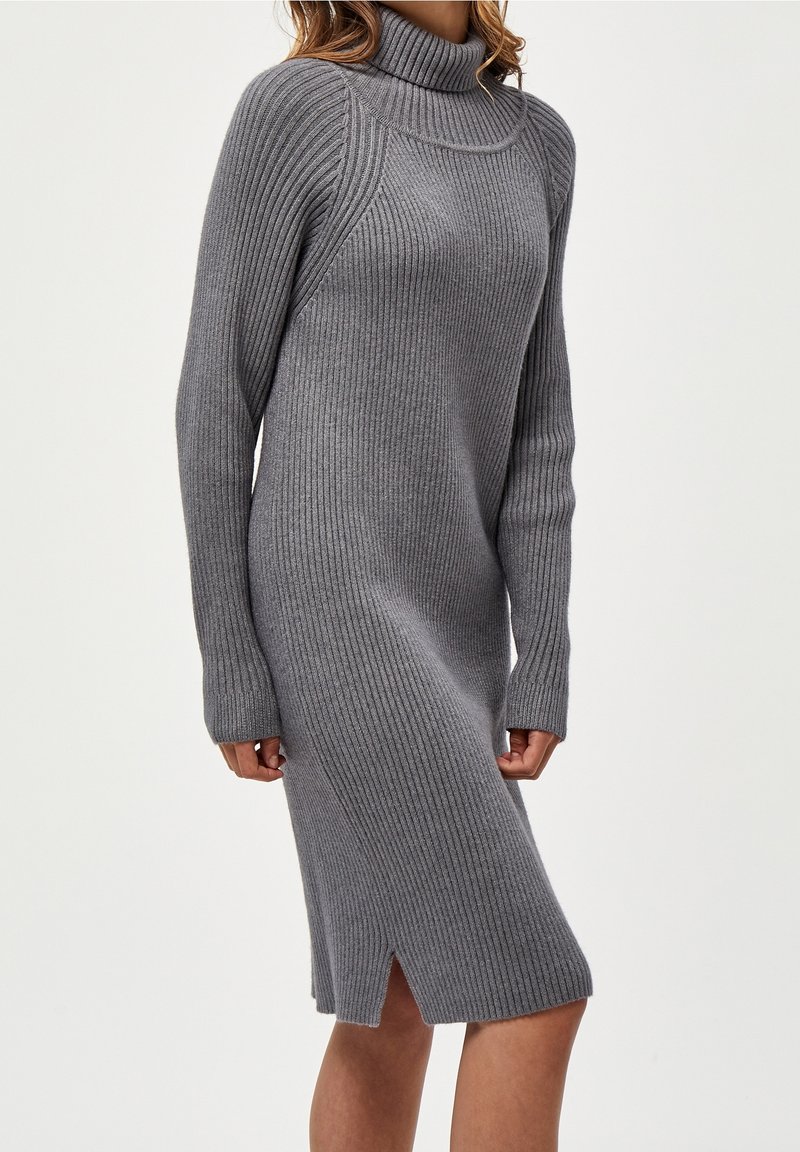 Femme portant une robe grise côtelée en maille, manches longues, longueur genou, col roulé et fente latérale, sur fond uni.