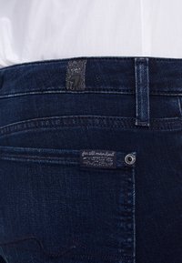 Jeans de mezclilla azul oscuro con una etiqueta de cintura texturizada en gris y una pequeña etiqueta de marca en el bolsillo trasero, que cuenta con un sutil detalle de costura.