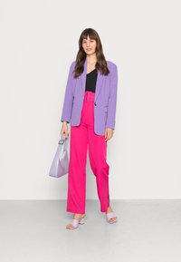 Blazer lilás sobre uma blusa preta, combinado com calças rosa de cintura alta. Acessorizado com uma bolsa lilás clara e sandálias a condizer.
