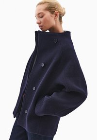 Granatowa kurtka z wełny z domieszką, z wysokim kołnierzem, dużymi guzikami i teksturowanym wykończeniem, charakteryzująca się luźnym krojem i bocznymi kieszeniami.