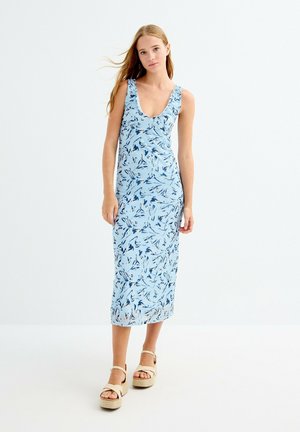 Joven mujer de pie con vestido midi azul sin mangas con estampado floral y sandalias beige de plataforma sobre fondo liso.