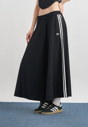 Kvinde iført sorte Adidas bukser med vide ben og hvide striber, sorte Adidas sneakers og hvide sokker på et blåt tæppe.
