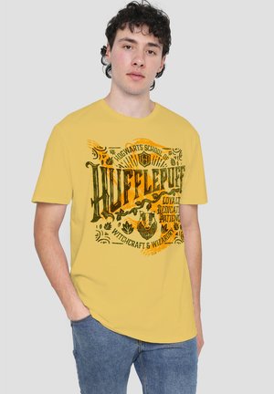 Young man wearing a yellow Hufflepuff t-shirt with black text and graphics, standing with one hand in pocket against a gray background.