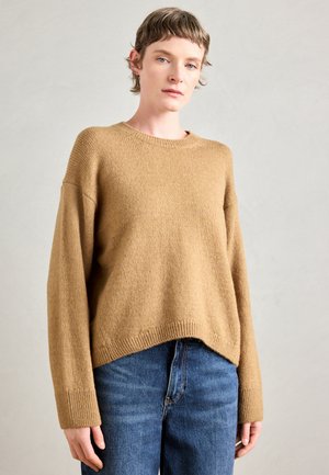 Brun strikkegenser med rund halslinning, senkede skuldre og lange ermer. Bærer over blå denimjeans, har en avslappet passform og ribbestrikkede detaljer.