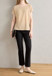 Camisa beige de manga corta con escote redondeado, combinada con pantalones negros ajustados y bailarinas beige claro, sosteniendo un bolso de mano negro.