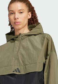 Olive green half-zip pullover jacket with hood, textured fabric, black lower section, and prominent black logo on the front.