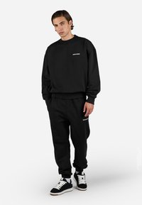 Sort sweatshirt og jogger-sæt, i bomuldsstof, afslappet pasform, ribbede manchetter, med et lille logo på brystet og låret, parret med sort-hvide sneakers.