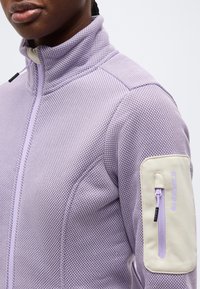 Άτομο που φοράει υφασμάτινο μωβ μπουφάν με φερμουάρ και τσέπη με φερμουάρ σε κρεμ χρώμα στον πάνω βραχίονα, μπροστά σε λευκό φόντο.