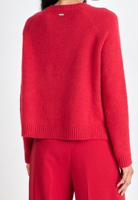 Pull rouge en maille avec ourlet et poignets côtelés, doté d'un col rond et d'un petit logo à l'arrière du col, confectionné en tissu doux.