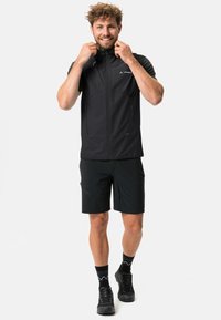 Giacca leggera nera con chiusura con zip, maniche corte e due tasche frontali. Indossata con pantaloni corti neri e calzature nere.