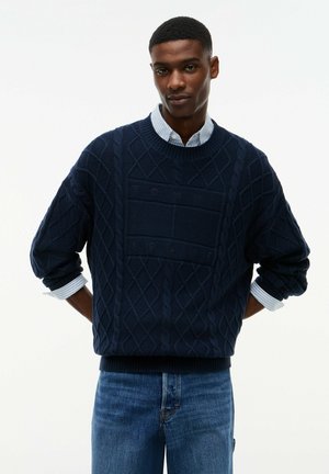 Junger Mann trägt einen dunkelblauen kabelgestrickten Pullover mit Muster und blaue Jeans, die Hände hinter dem Rücken, stehend vor einem einfarbigen weißen Hintergrund.