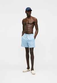 Särgi mitte kandnud mees, kes kannab helesiniseid paelaga lühikesi pükse, tumesinist nokamütsi ja valgeid slaiprisid, seisab käed taskutes ühevärvilise tausta ees.