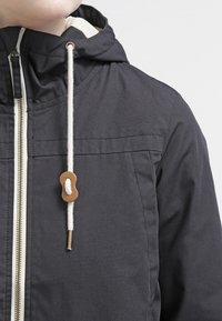 Svart huoodjacka med dragkedja framtill, krämfärgad foder och snörning med en brun träknapp. Strukturerad tyg med sömnadsdetaljer.