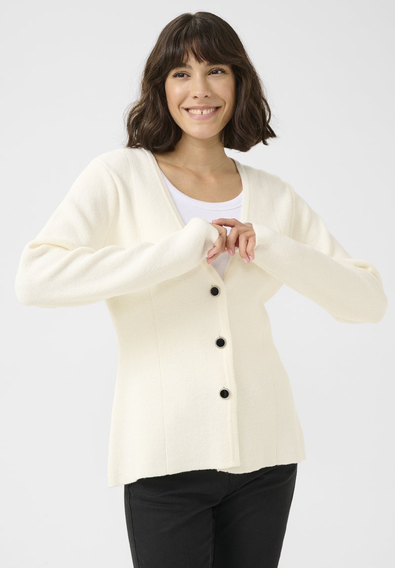 Cardigan blanc en tricot avec un col en V, des boutons à l'avant et des manches longues. Tissu texturé et silhouette ajustée.