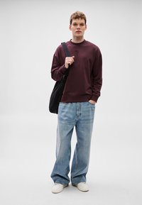 Bordó pulóver kerek nyakkivágással, világoskék, bő szárú farmerrel és fehér tornacipővel párosítva. Fekete válltáska a vállán.