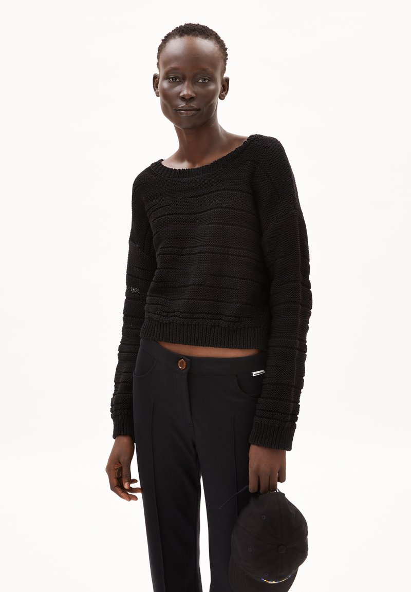 Schwarzer gestrickter, kurzer Pullover mit einem strukturierten Streifenmuster, kombiniert mit schwarzen Hosen, die einen braunen Knopf und Taschen haben.